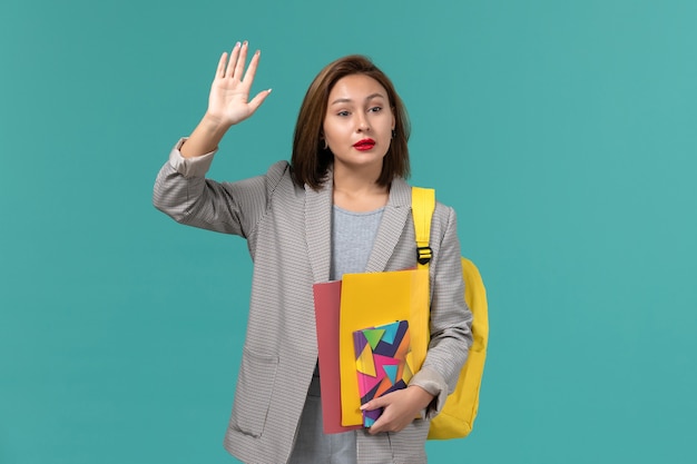 Front view of female student in grey jacket wearing yellow backpack holding files and copybook waving on blue wall