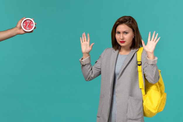 Front view of female student in grey jacket wearing yellow backpack on light-blue wall