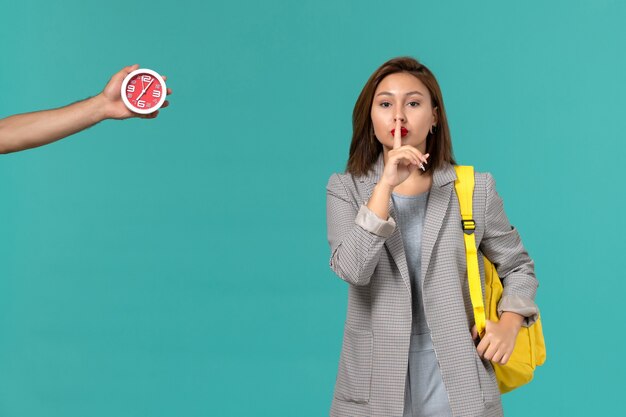 Front view of female student in grey jacket wearing yellow backpack on light blue wall