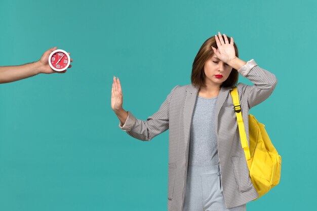 Front view of female student in grey jacket wearing yellow backpack on the light-blue wall