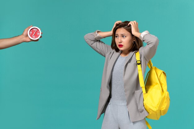 Front view of female student in grey jacket wearing yellow backpack posing with confused expression on light blue wall