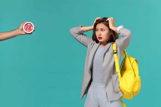 Front view of female student in grey jacket wearing yellow backpack posing with confused expression on light blue wall