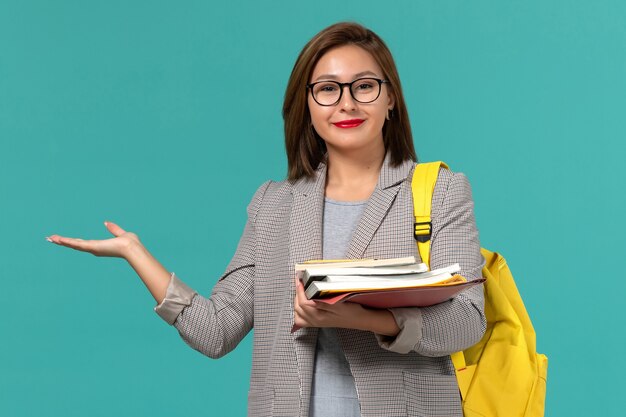Front view of female student in grey jacket yellow backpack holding books on light blue wall