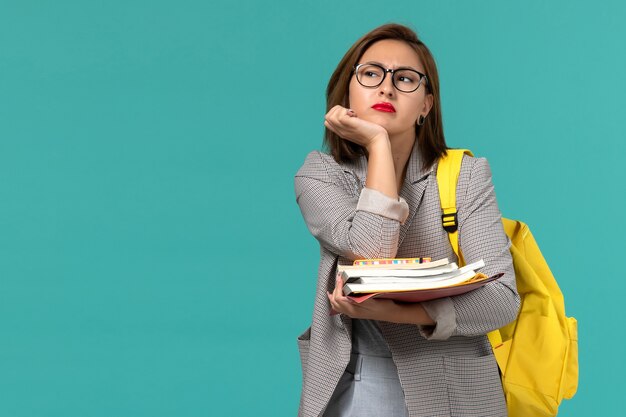 Front view of female student in grey jacket yellow backpack holding books thinking on blue wall