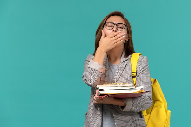 青い壁にあくびをしている本を保持している灰色のジャケット黄色のバックパックの女子学生の正面図