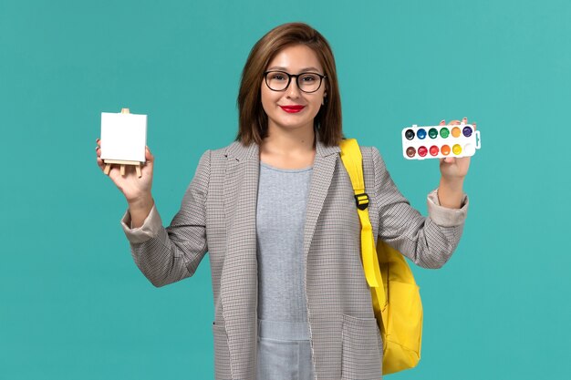 Front view of female student in grey jacket yellow backpack holding paints and easel on blue wall