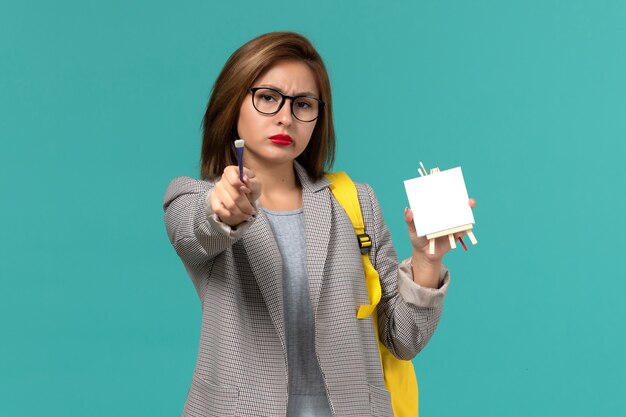 Front view of female student in grey jacket yellow backpack holding tassel and easel on blue wall