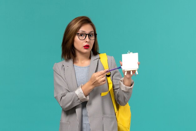 Front view of female student in grey jacket yellow backpack holding tassels and easel on blue wall