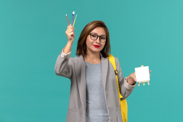 Front view of female student in grey jacket yellow backpack holding tassels and easel on the blue wall