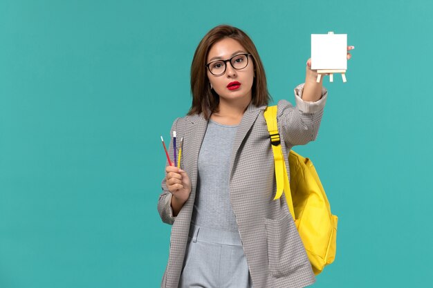 Front view of female student in grey jacket yellow backpack holding tassels and easel on light blue wall