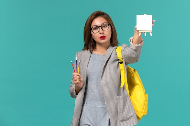 Front view of female student in grey jacket yellow backpack holding tassels and easel on light blue wall