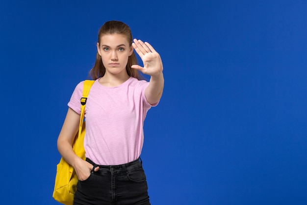 Front view of female student in pink t-shirt with yellow backpack on blue wall