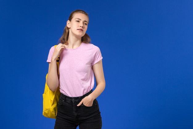 Front view of female student in pink t-shirt with yellow backpack on blue wall