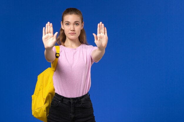 Front view of female student in pink t-shirt with yellow backpack on the blue wall