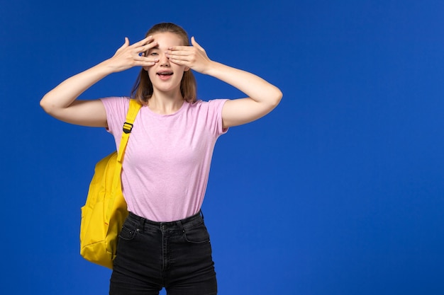 Front view of female student in pink t-shirt with yellow backpack closing her eyes on light blue wall