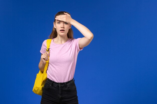 Front view of female student in pink t-shirt with yellow backpack confused expression on the blue wall