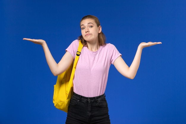 水色の壁に混乱している黄色のバックパックとピンクのTシャツの女子学生の正面図