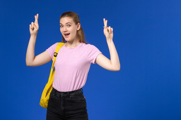 Вид спереди студентки в розовой футболке с желтым рюкзаком, скрестив пальцы на синей стене