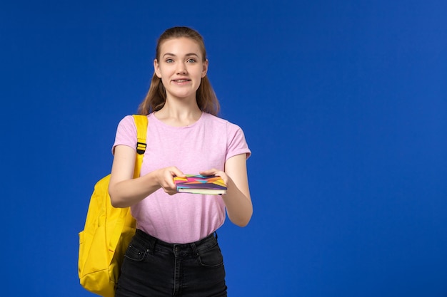 青い壁にコピーブックを保持している黄色のバックパックとピンクのTシャツの女子学生の正面図