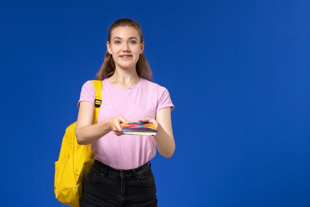 Вид спереди студентки в розовой футболке с желтым рюкзаком, держащей тетрадь на синей стене