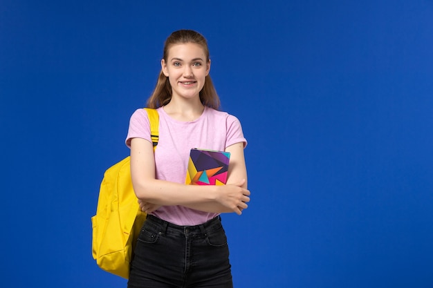 青い壁にコピーブックを保持している黄色のバックパックとピンクのTシャツの女子学生の正面図