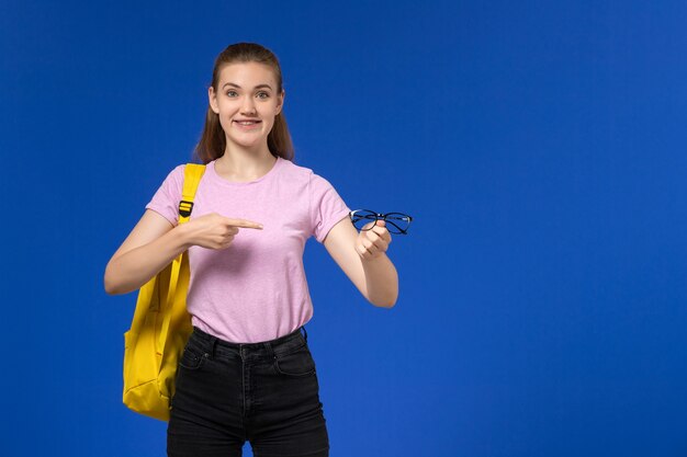 青い壁に光学サングラスをかけた黄色のバックパックが付いたピンクのTシャツを着た女子学生の正面図