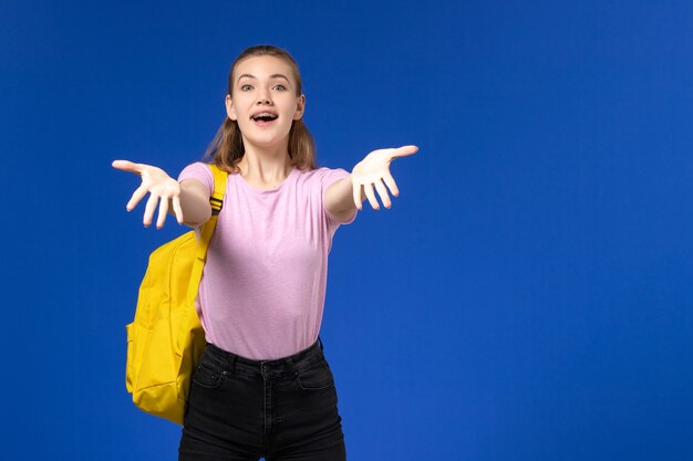 Front view of female student in pink t-shirt with yellow backpack on light-blue wall