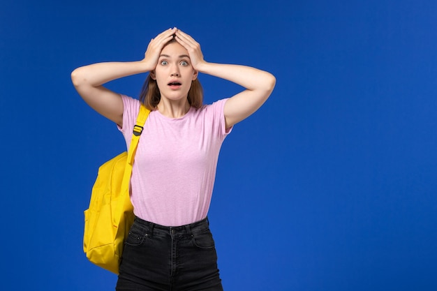 Front view of female student in pink t-shirt with yellow backpack on light blue wall