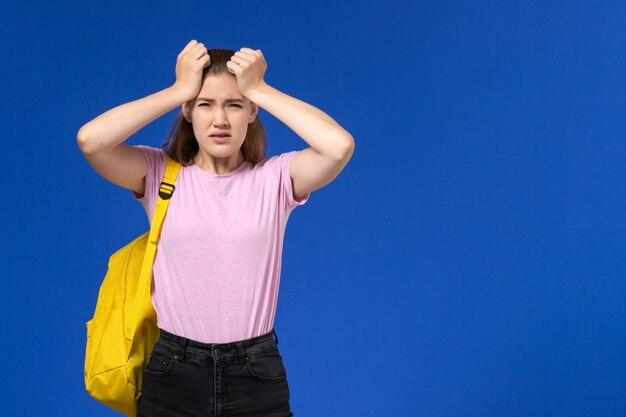 Front view of female student in pink t-shirt with yellow backpack on the light blue wall