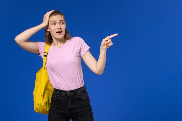 水色の壁に黄色のバックパックを指しているピンクのTシャツの女子学生の正面図