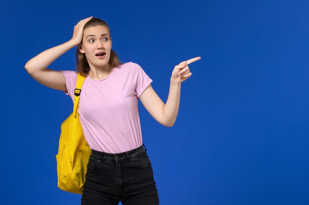 Front view of female student in pink t-shirt with yellow backpack pointing out on light-blue wall