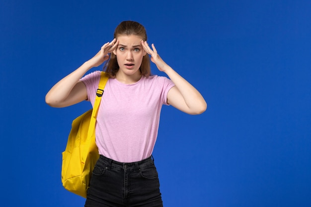 Вид спереди студентки в розовой футболке с желтым рюкзаком, позирующей на синей стене