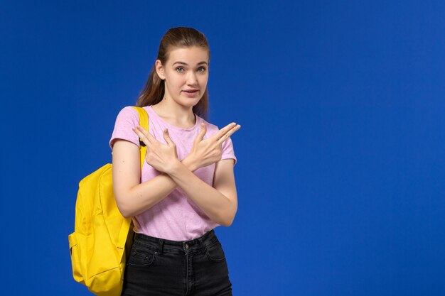 Front view of female student in pink t-shirt with yellow backpack posing on blue wall