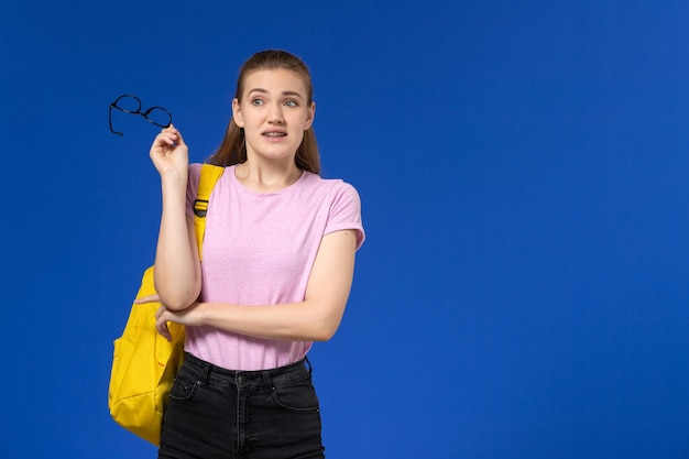 Front view of female student in pink t-shirt with yellow backpack posing on the blue wall
