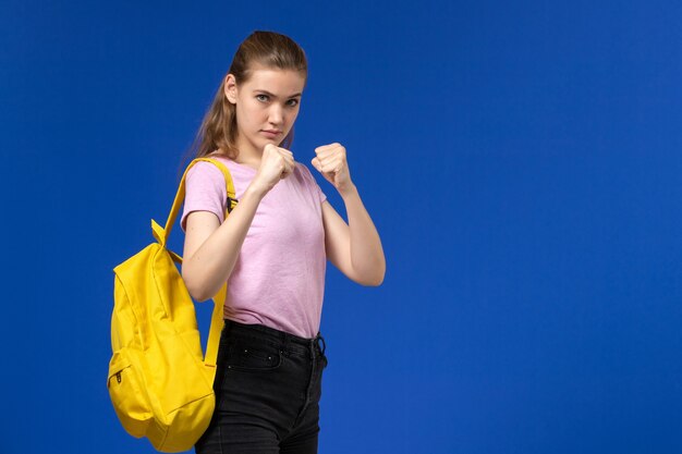 Front view of female student in pink t-shirt with yellow backpack posing in boxing stand on blue wall