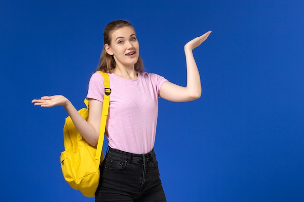 Vista frontale della studentessa in maglietta rosa con zaino giallo in posa sulla parete azzurra