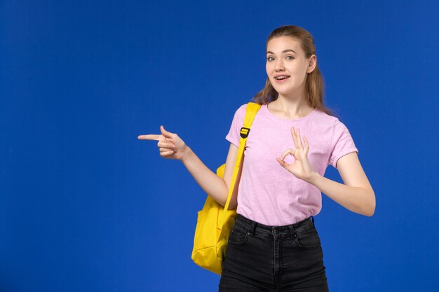 水色の壁にポーズをとって黄色のバックパックとピンクのTシャツの女子学生の正面図