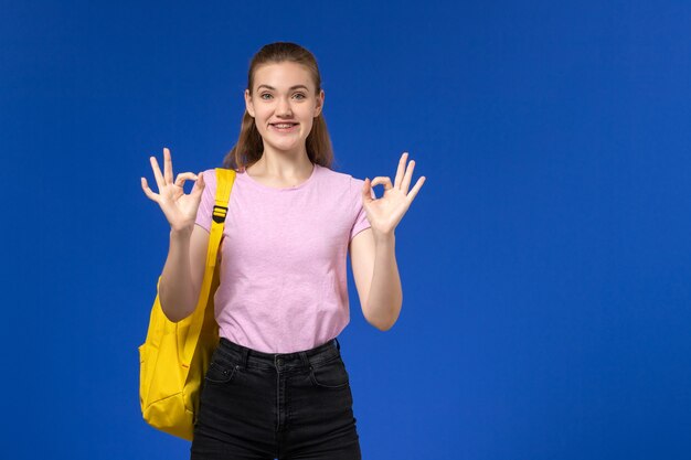 Front view of female student in pink t-shirt with yellow backpack posing and smiling on blue wall