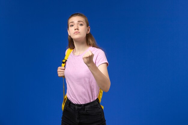 Front view of female student in pink t-shirt with yellow backpack posing and threatening on the light-blue wall