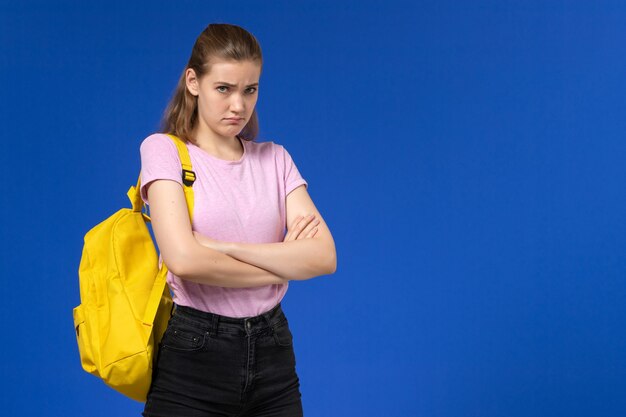 Front view of female student in pink t-shirt with yellow backpack posing with mad expression on light blue wall