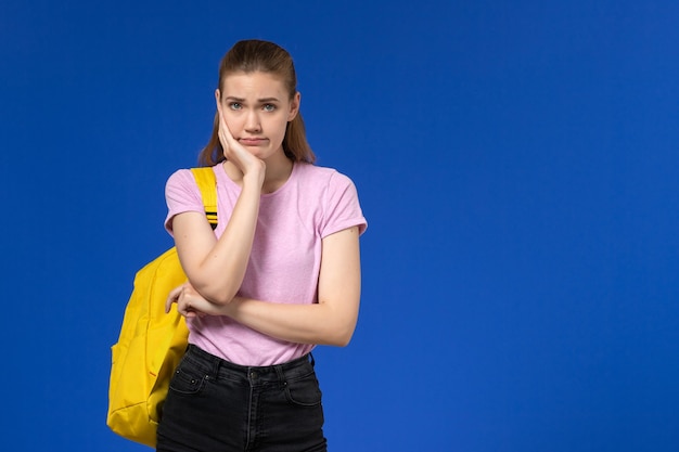 파란색 벽에 스트레스 표정으로 포즈를 취하는 노란색 배낭과 분홍색 티셔츠에 여성 학생의 전면보기