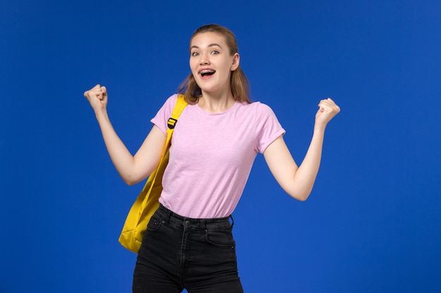 Вид спереди студентки в розовой футболке с желтым рюкзаком, радующейся на голубой стене