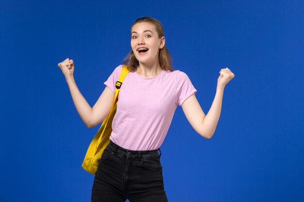 Front view of female student in pink t-shirt with yellow backpack rejoicing on light-blue wall