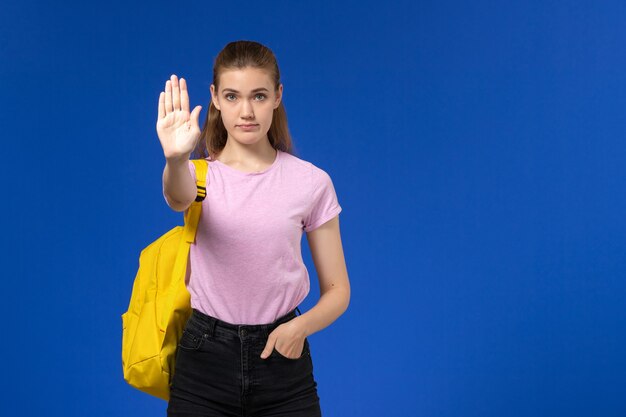 Front view of female student in pink t-shirt with yellow backpack showing stop sign on blue wall