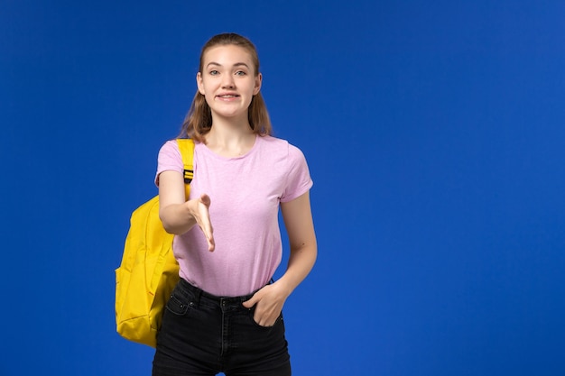 Вид спереди студентки в розовой футболке с желтым рюкзаком, улыбающейся на голубой стене