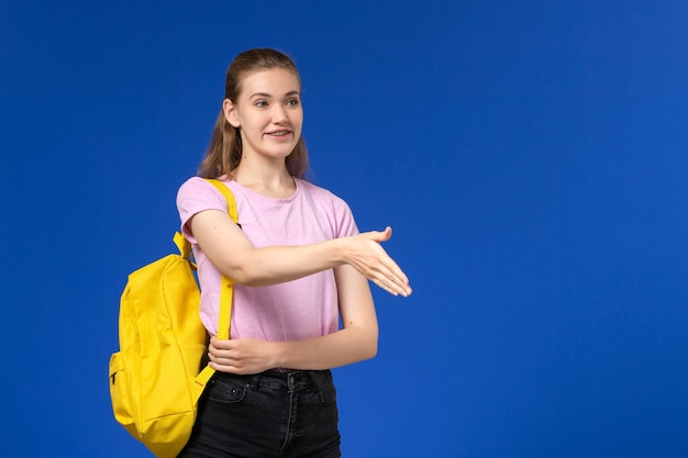 青い壁に笑顔と手を振って黄色のバックパックとピンクのTシャツの女子学生の正面図