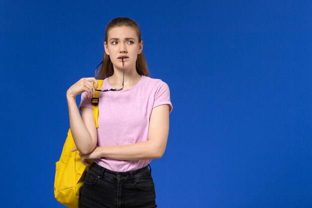 Вид спереди студентки в розовой футболке с желтым рюкзаком, думая на синей стене