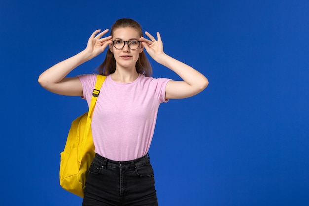 Вид спереди студентки в розовой футболке с желтым рюкзаком в оптических очках на синей стене