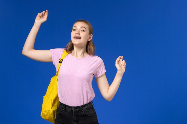 Front view of female student in pink t-shirt with yellow backpack yawning on light blue wall
