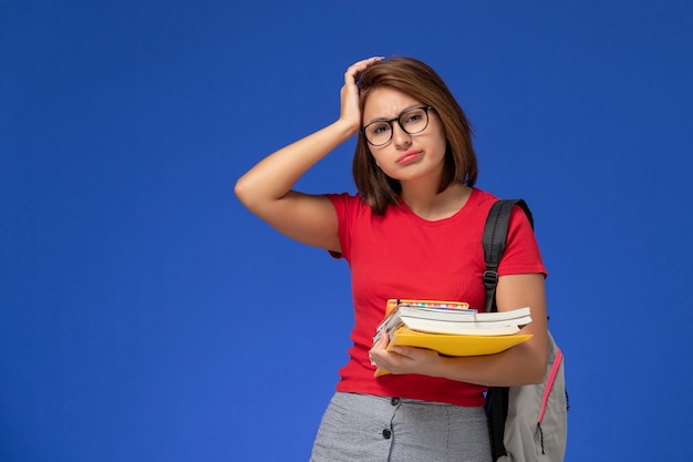 Вид спереди студентки в красной рубашке с рюкзаком, держащим книги и файлы на голубой стене